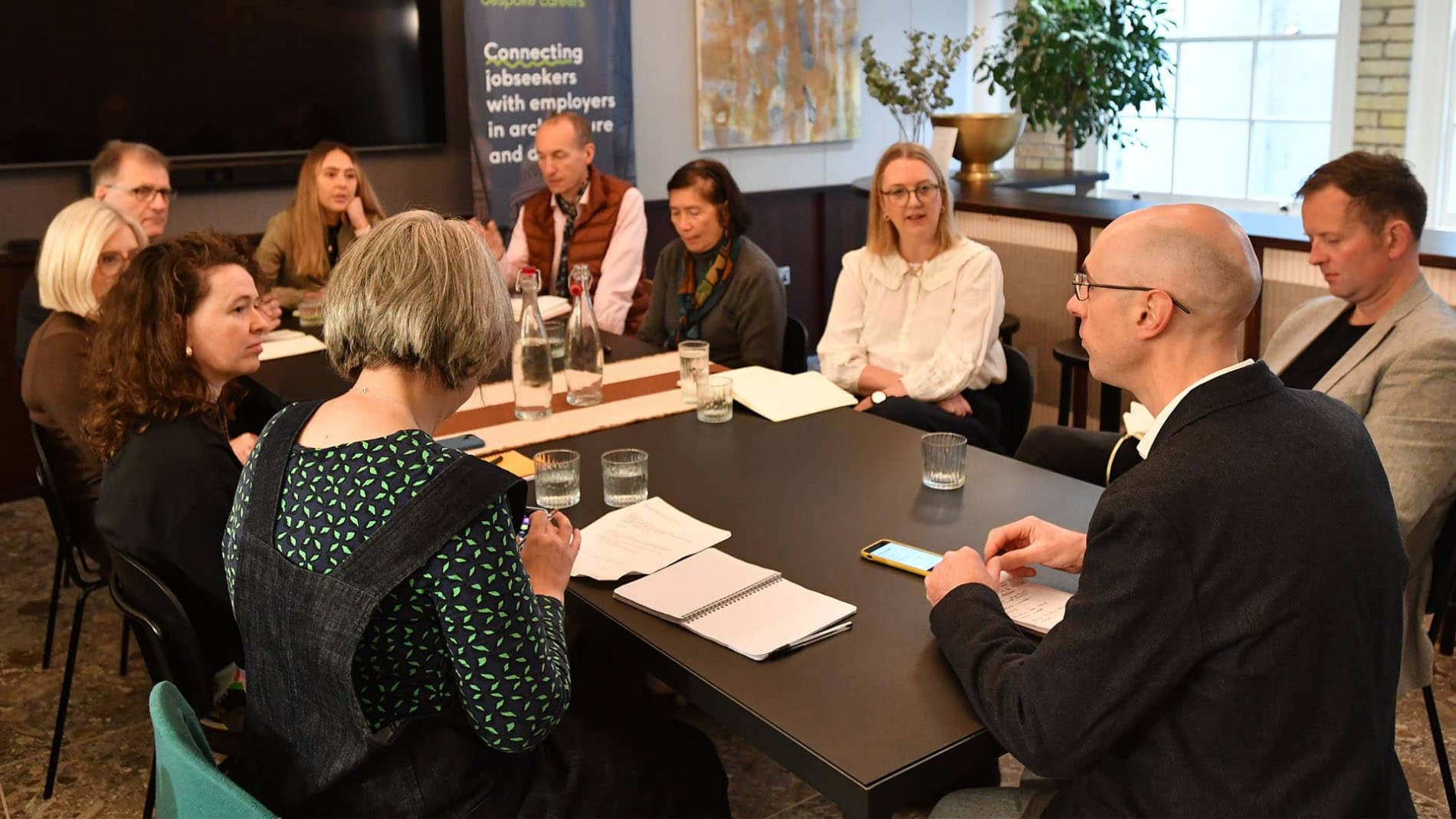 Business meeting with diverse professionals seated around a conference table. A banner in the background mentions connecting jobseekers with employers. Participants have notepads and water glasses on the table.