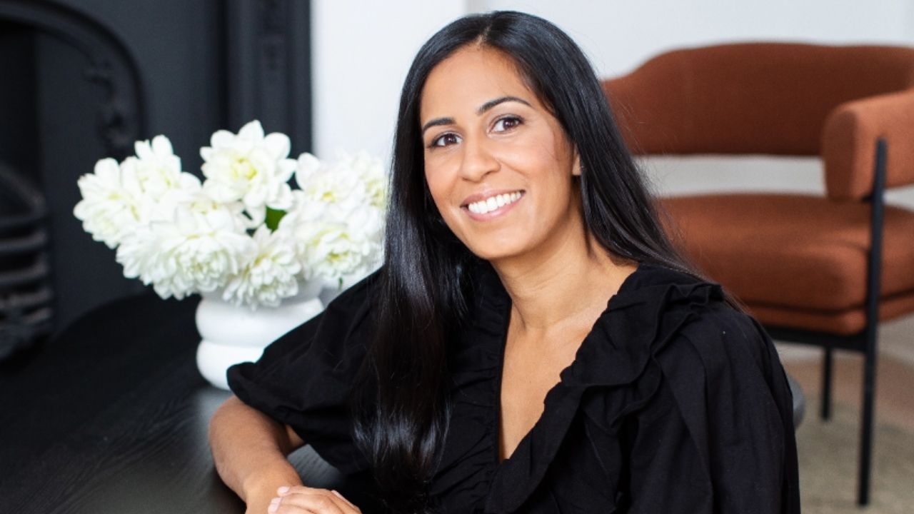 A profile of Nikita Morell. She is smiling at the camera. She is sitting on the floor in a lounge. There are flowers in the background.