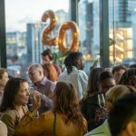 A party in London. there are lots of people drinking champagne. There are balloons spelling out "20"