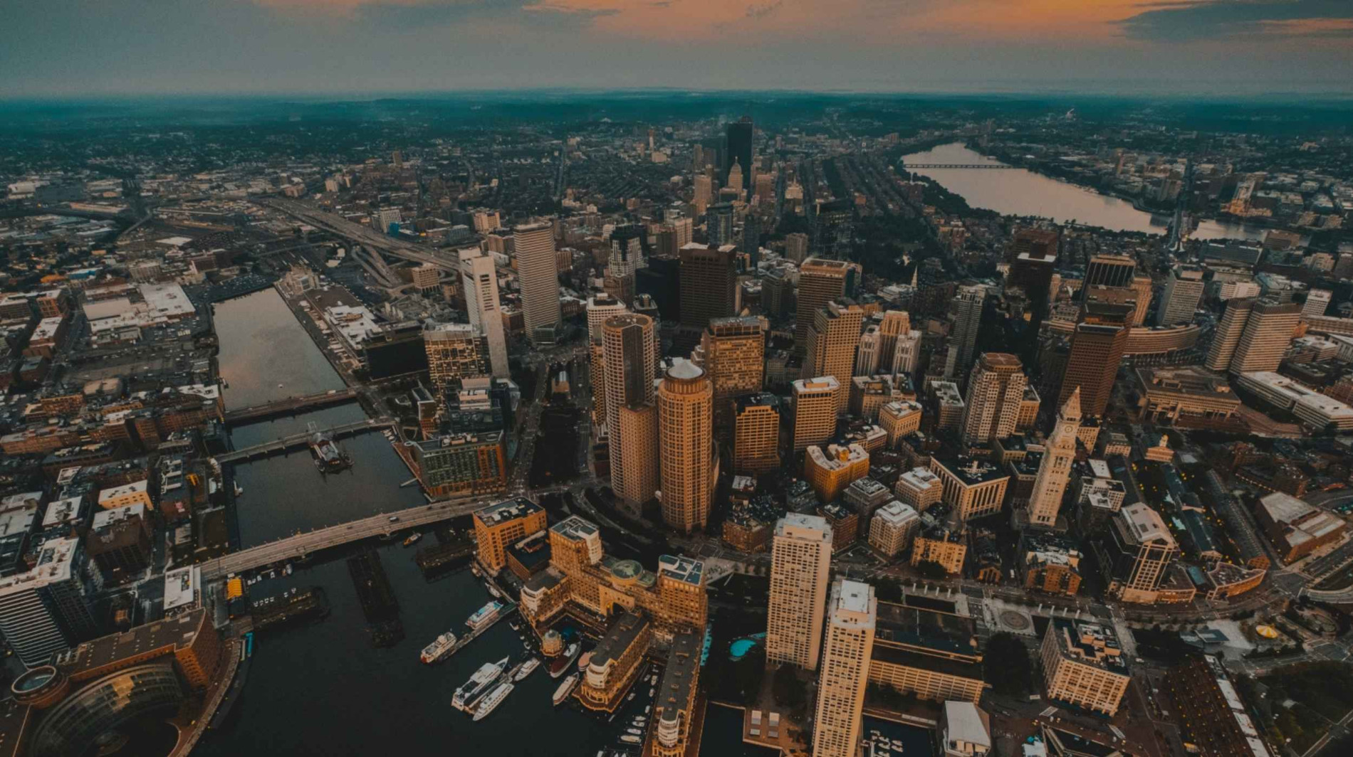 Aerial view of downtown Boston at sunset, showing the harbor, bridges, and skyscrapers along the waterfront with the Charles River in the distance.