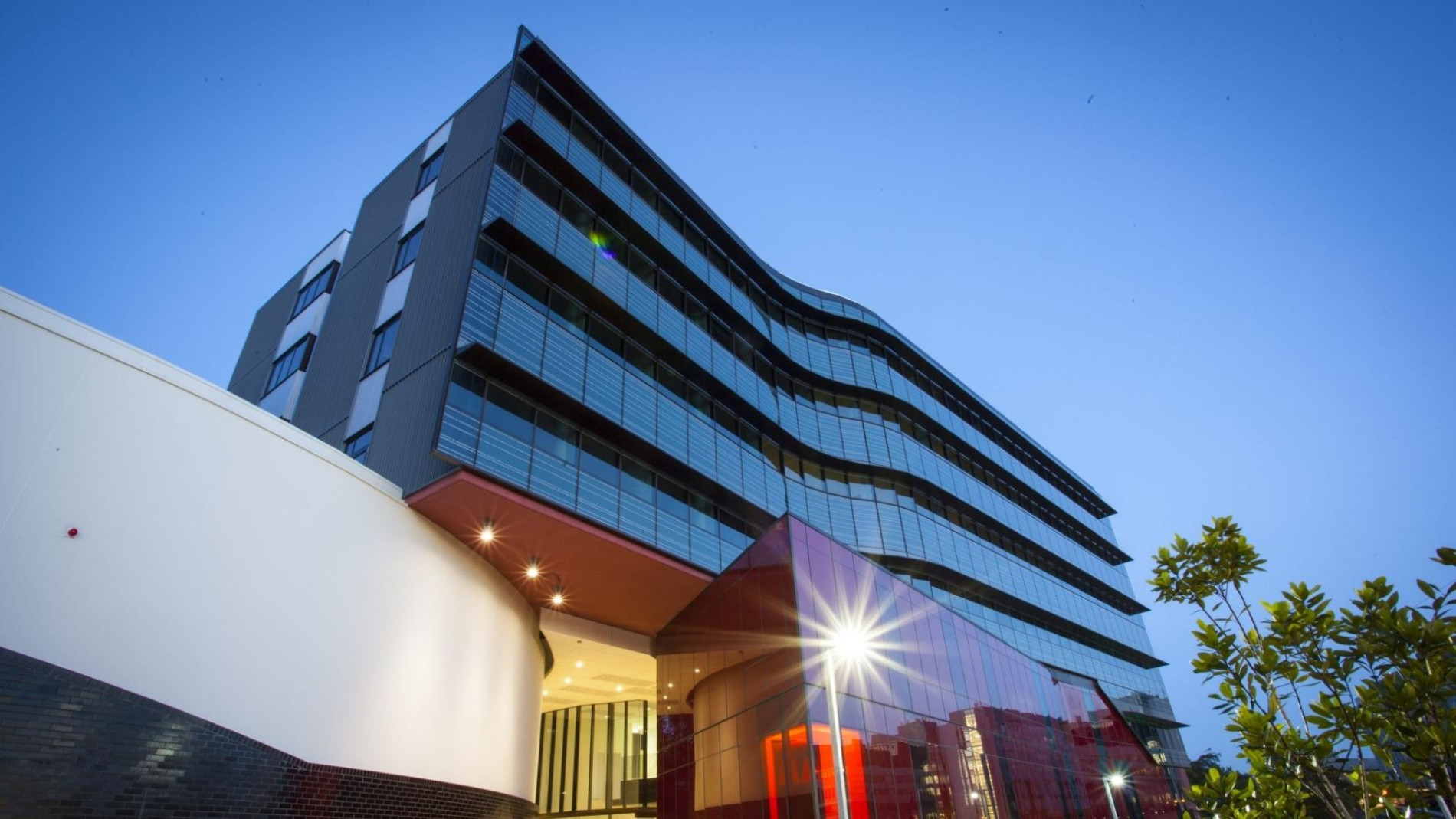 Modern commercial building with a sleek, curved glass façade, angular red accents, and black-and-white finishes, set against a twilight sky and surrounded by landscaping.