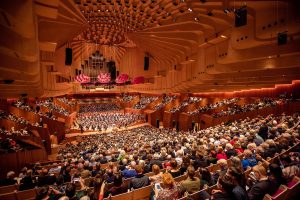 Interior of Sydney Opera House concert hall during a performance, showing full orchestra on stage and packed audience seating, with distinctive wood-paneled architecture and pink suspended art installation