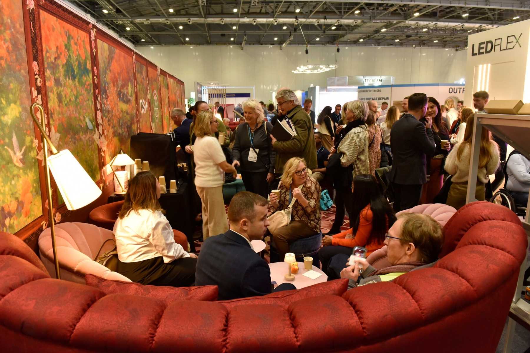 Trade show lounge area with crowded networking session. Features a large red curved sofa, decorative wall murals, and table lamps. Attendees are standing and sitting, engaged in conversation, with some holding beverages. LEDFLEX signage visible in background.