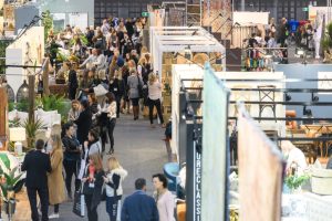 Crowded aisle at a decor and design trade show with attendees browsing white exhibition booths.