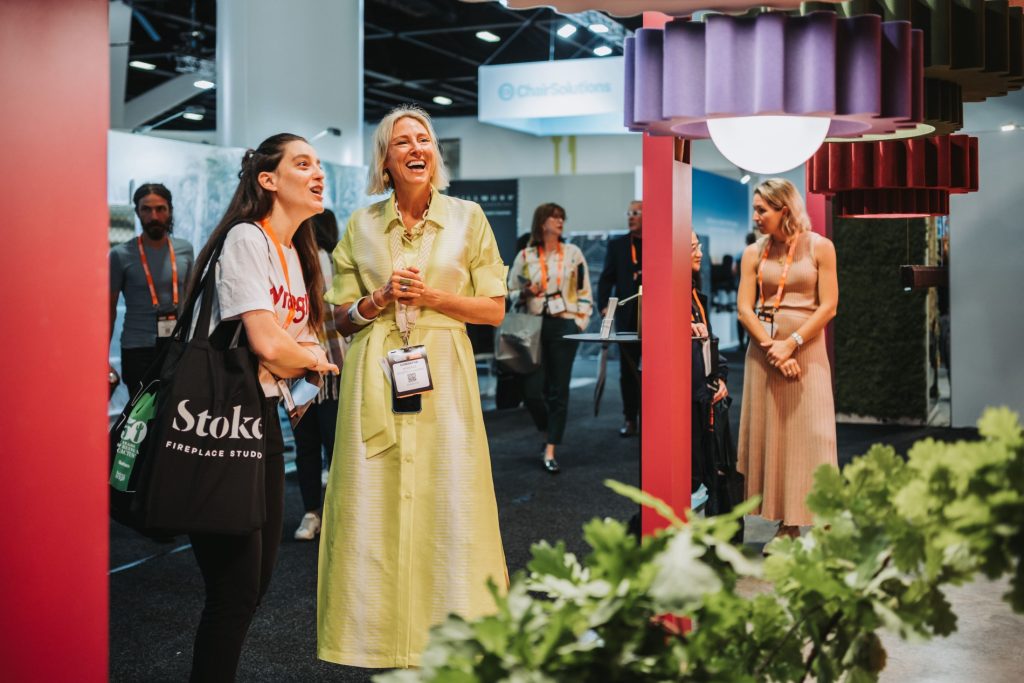 Two women laughing together at a trade show or convention. One wears a casual white t-shirt and carries a Stoke Fireplace Studio tote bag, while the other wears a yellow dress. Both are wearing event lanyards.