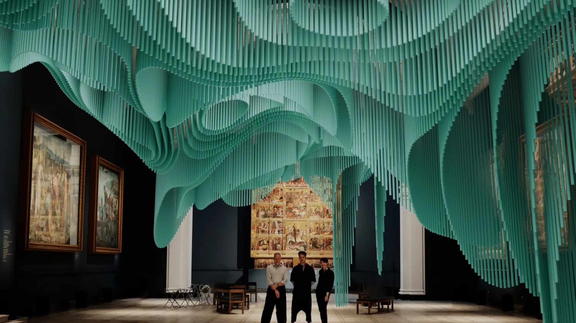 Three people stand beneath a striking turquoise installation of cascading vertical elements, displayed in a gallery featuring historic artworks and intricate flooring.