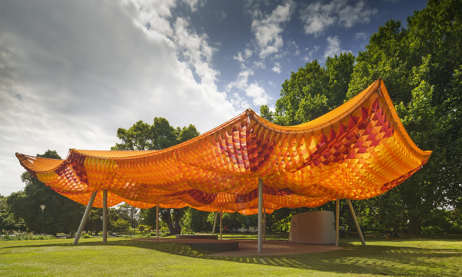 Vibrant pavilion structure featuring a dynamic orange and red patterned canopy, supported by sleek metal columns, set in a lush green park with a dramatic sky in the background