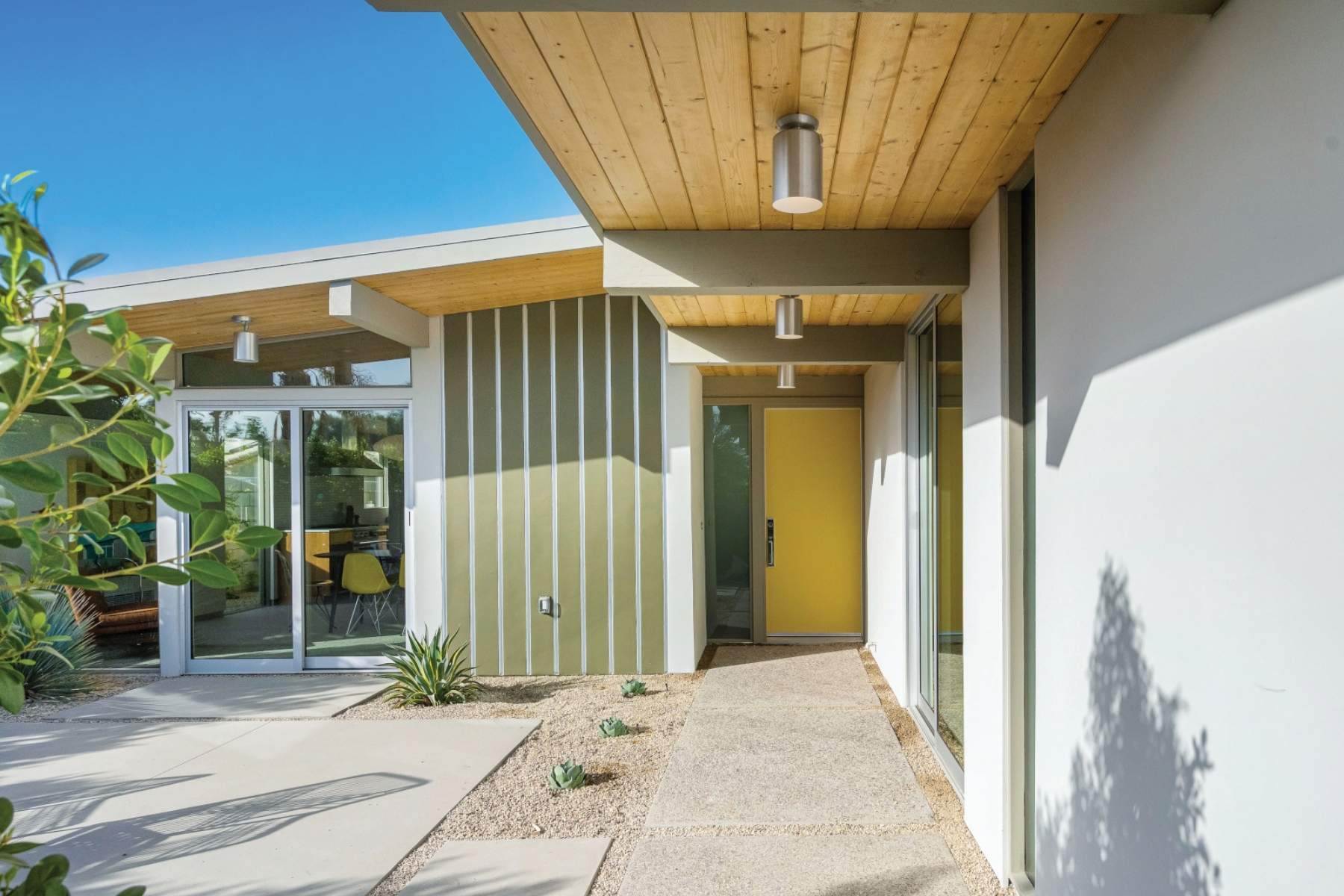 Modern mid-century style home entrance with yellow door, wood-paneled ceiling, and desert landscaping. Features floor-to-ceiling windows and a covered walkway.