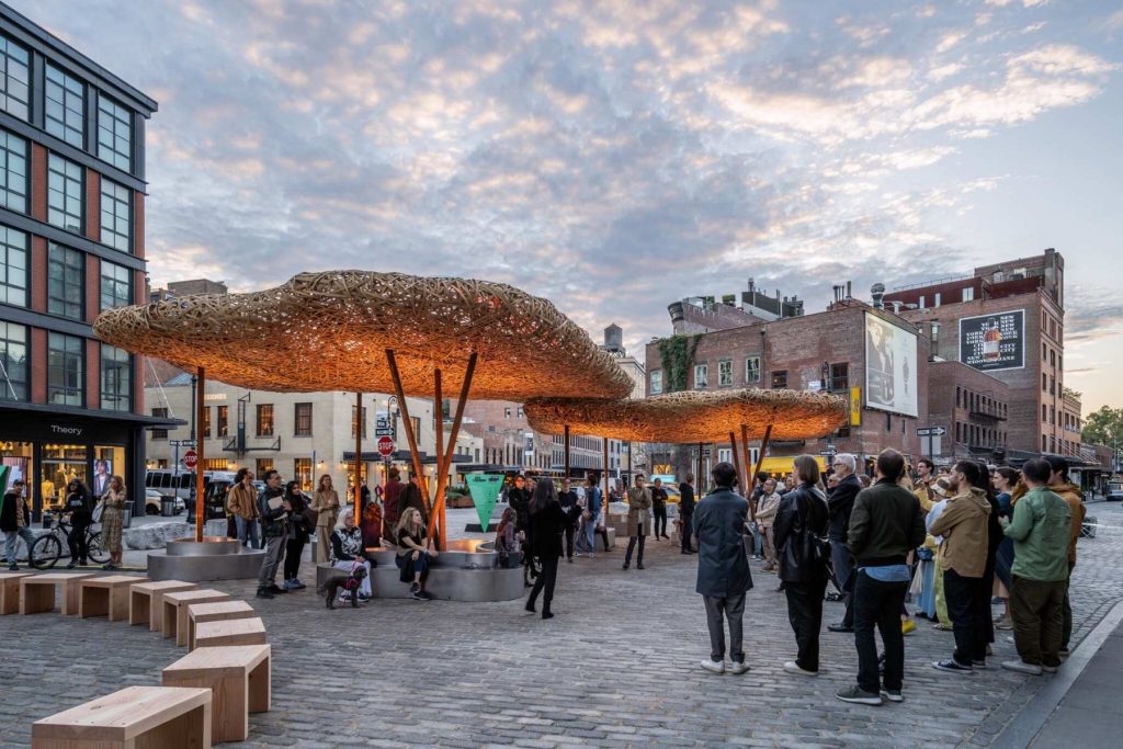 New York City's Meatpacking District, featuring the distinctive woven canopy art installation "Head Up" by Talley Fisher. The large, illuminated copper-colored structures hover over a cobblestone plaza where people are gathered in the evening.