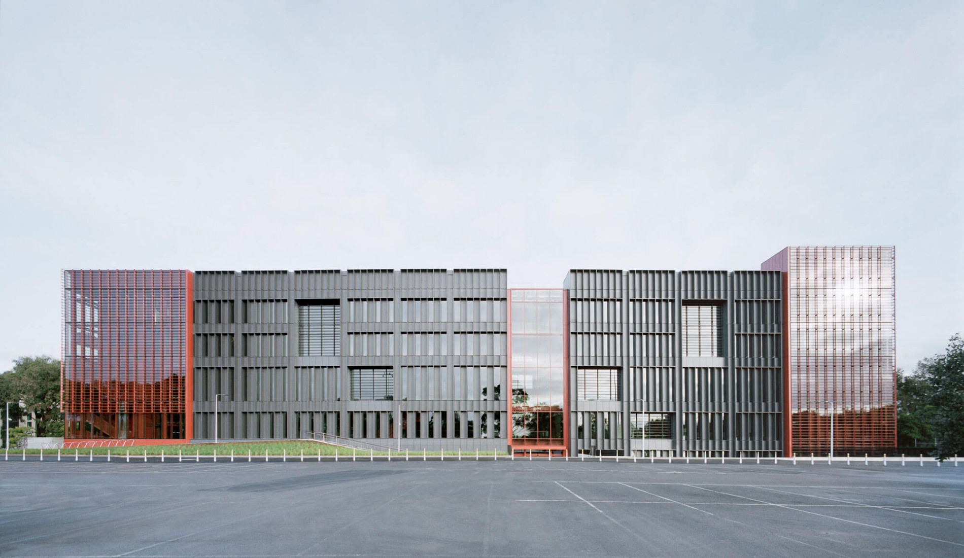 A modern office building with a distinctive facade featuring vertical metal louvers in gray and rust-red colors, with a large parking lot in the foreground