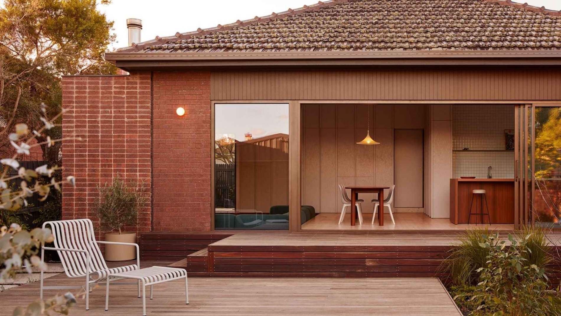 Exterior view of a modern Australian home featuring a warm brick façade, timber decking, and an inviting indoor-outdoor connection, with contemporary outdoor furniture and lush greenery enhancing the space."