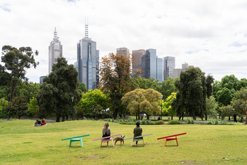 A peaceful urban park scene featuring colorful, minimalist benches against a backdrop of Melbourne's city skyline. A couple sits on separate benches, enjoying the greenery and calm surroundings.