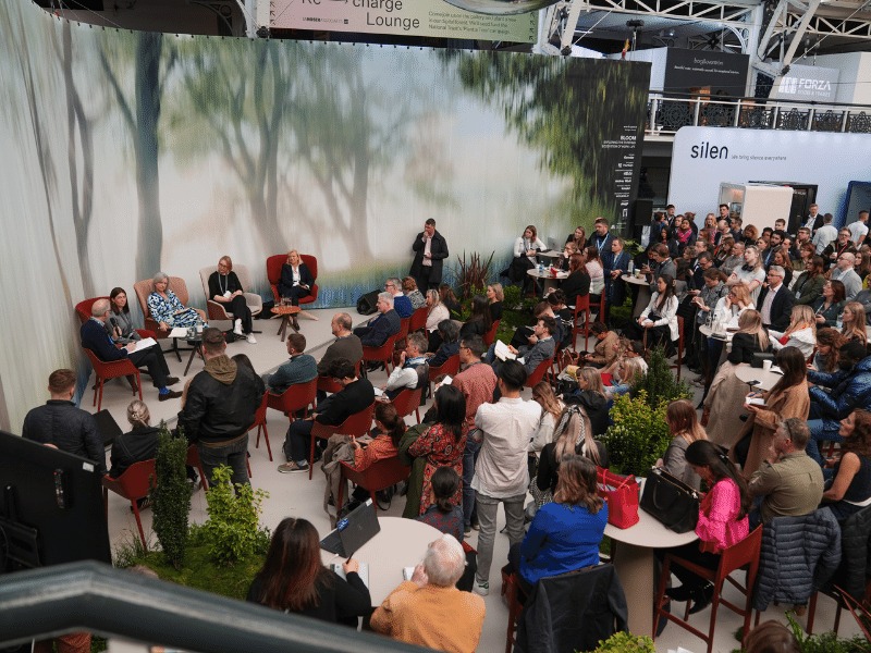 Large crows listens in to panel speaking. there is a striking background behind the speakers featuring an abstract tree on a screen