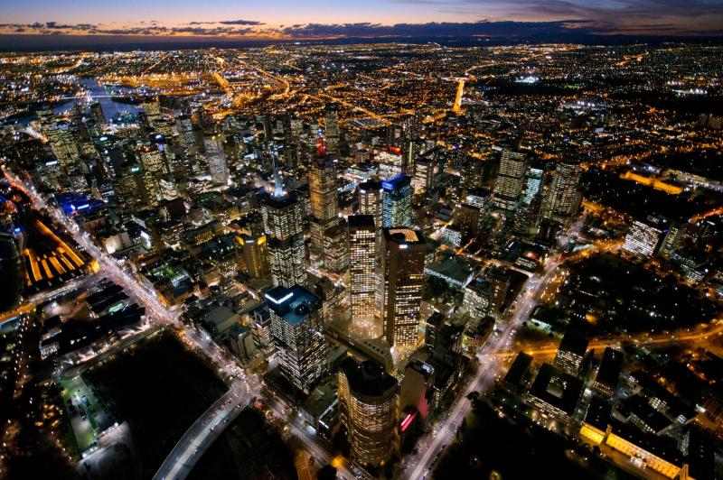 An aerial view of a bustling city skyline at dusk, with tall skyscrapers lit up against a darkening sky. Streets are lined with glowing streetlights, and the city extends into the distance, highlighting a vibrant urban landscape."