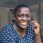 Profile of Ayo Abbas. She has short dark hair and is smiling at the camera. The background is the London skyline.