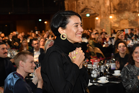 A woman in a black outfit stands and smiles with clasped hands at a formal awards event, surrounded by an applauding audience. The venue has warm lighting and rustic exposed walls, with tables set for a banquet.