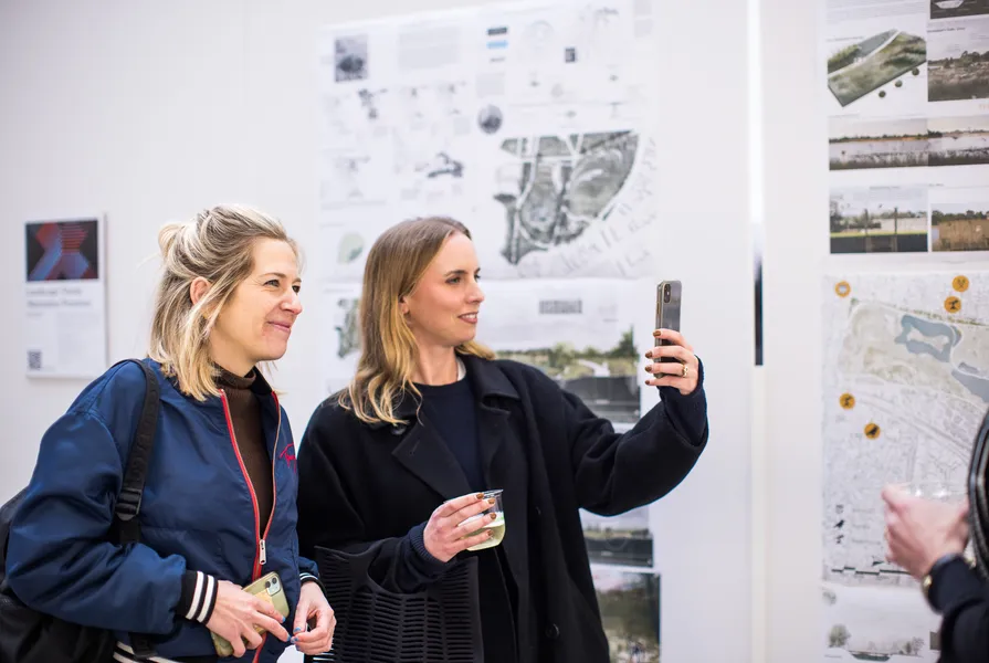 Two women enjoying a design exhibition, one taking a selfie while holding a drink, with architectural and landscape design boards displayed in the background.