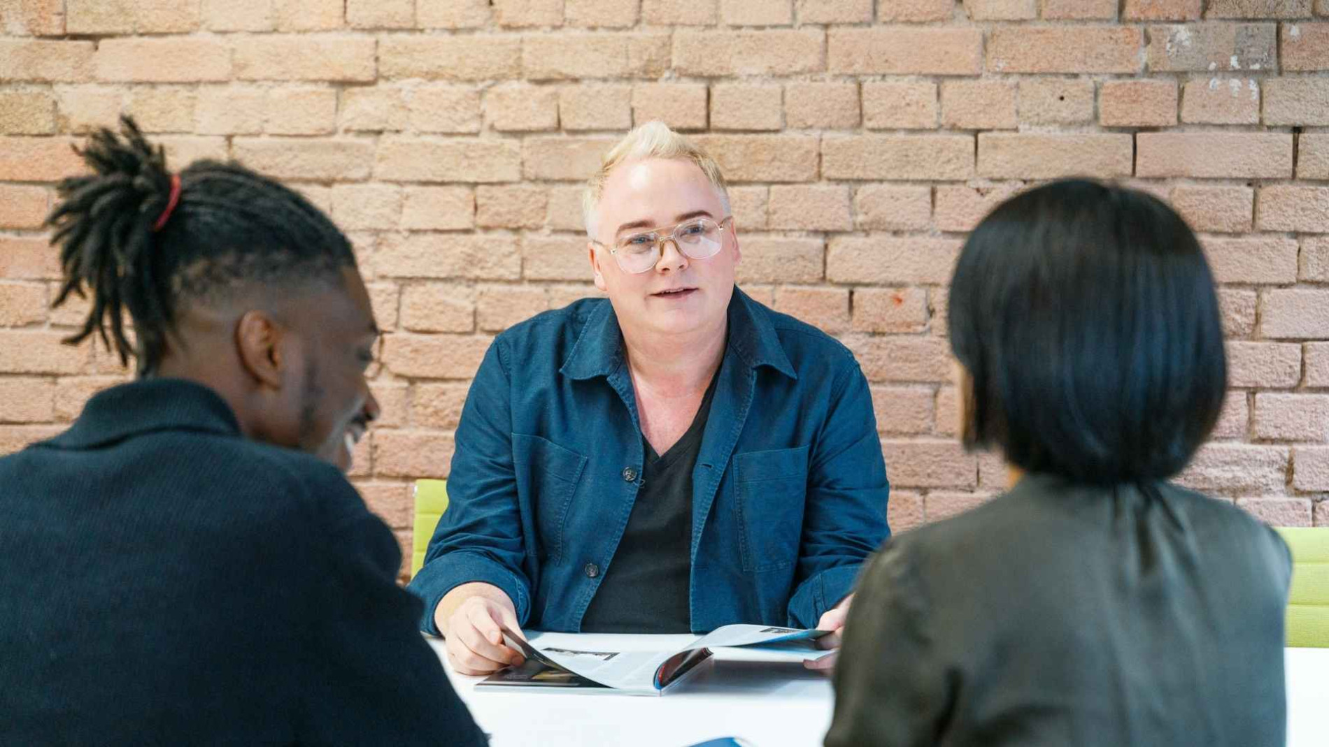 This image shows three people in a meeting - a white-haired man in a blue jacket, a man with dreadlocks, and a woman with dark hair. They appear to be having a discussion around a table.