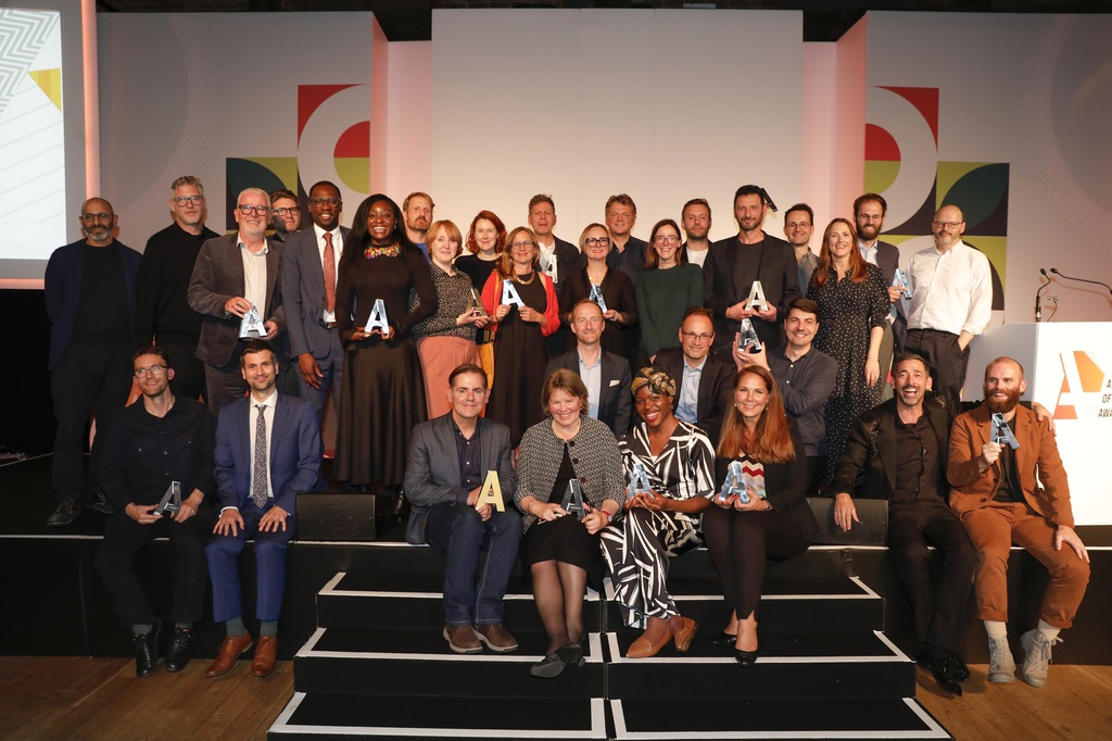 Group photo of winners and participants at the Architect of the Year Awards, hosted by Building Design. The image shows a diverse group of architects holding trophies, celebrating their achievements on a brightly lit stage with a colourful backdrop.