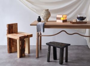  Handcrafted furniture and decor pieces, featuring a rustic wood chair, a blackened wood stool, a minimalist wooden table with curved details, and an assortment of artisanal ceramics and a stained glass lamp, styled against a neutral backdrop.