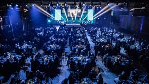 RIBA Stirling Prize 2024 awards ceremony with attendees seated at round tables in a dimly lit venue, showcasing a vibrant stage and dramatic lighting effects.