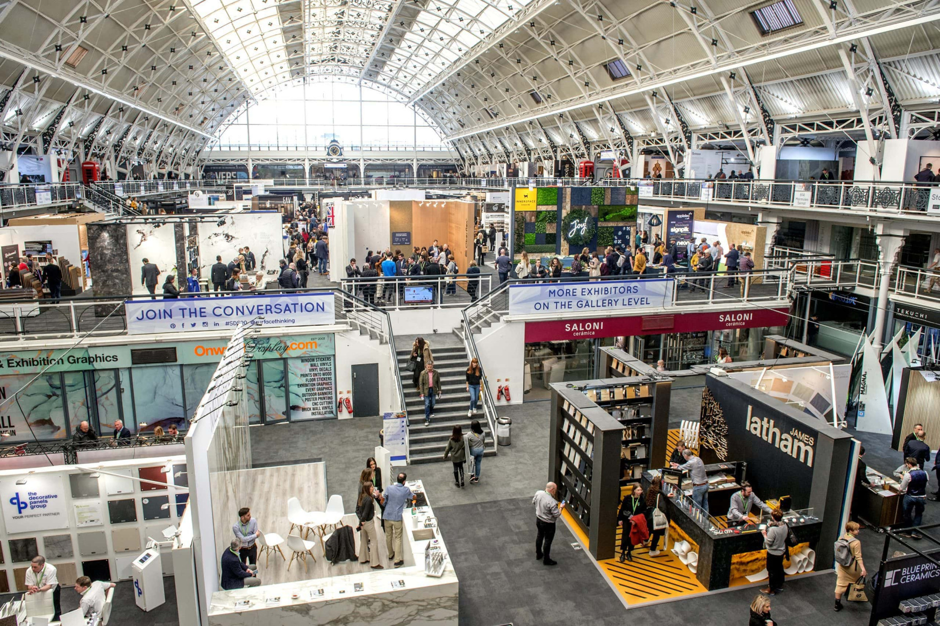 Interior view of the Business Design Centre during an event, showcasing multiple exhibition booths, vibrant displays, and attendees engaging in networking and discussions under the centre's iconic arched roof."