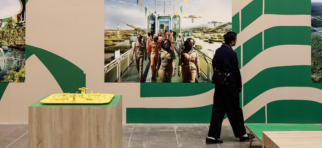 Exhibition display featuring a vibrant model in yellow on a wooden stand, with a large photograph of individuals in uniform walking on a futuristic bridge surrounded by green and natural landscapes, against a green and beige graphic wall backdrop."