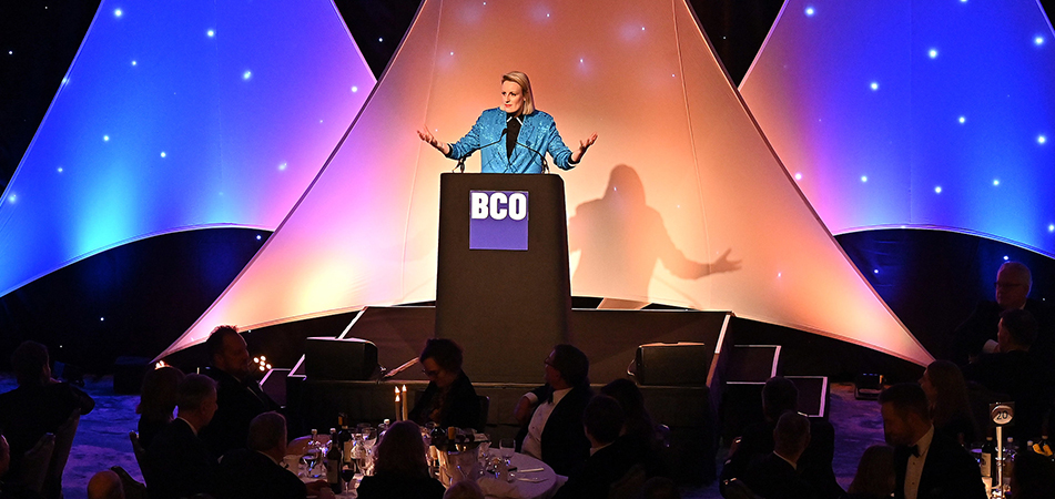 Speaker addressing the audience at a BCO Annual Dinner event, standing at a podium with a blue and purple-lit stage backdrop.