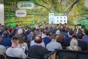 Panel discussion at the Sustainability Stage during the London Build Expo, featuring speakers discussing sustainability leadership. The audience is engaged, wearing blue headphones for a silent conference experience, with a backdrop of a large green tree and event sponsor logos including Amazon.