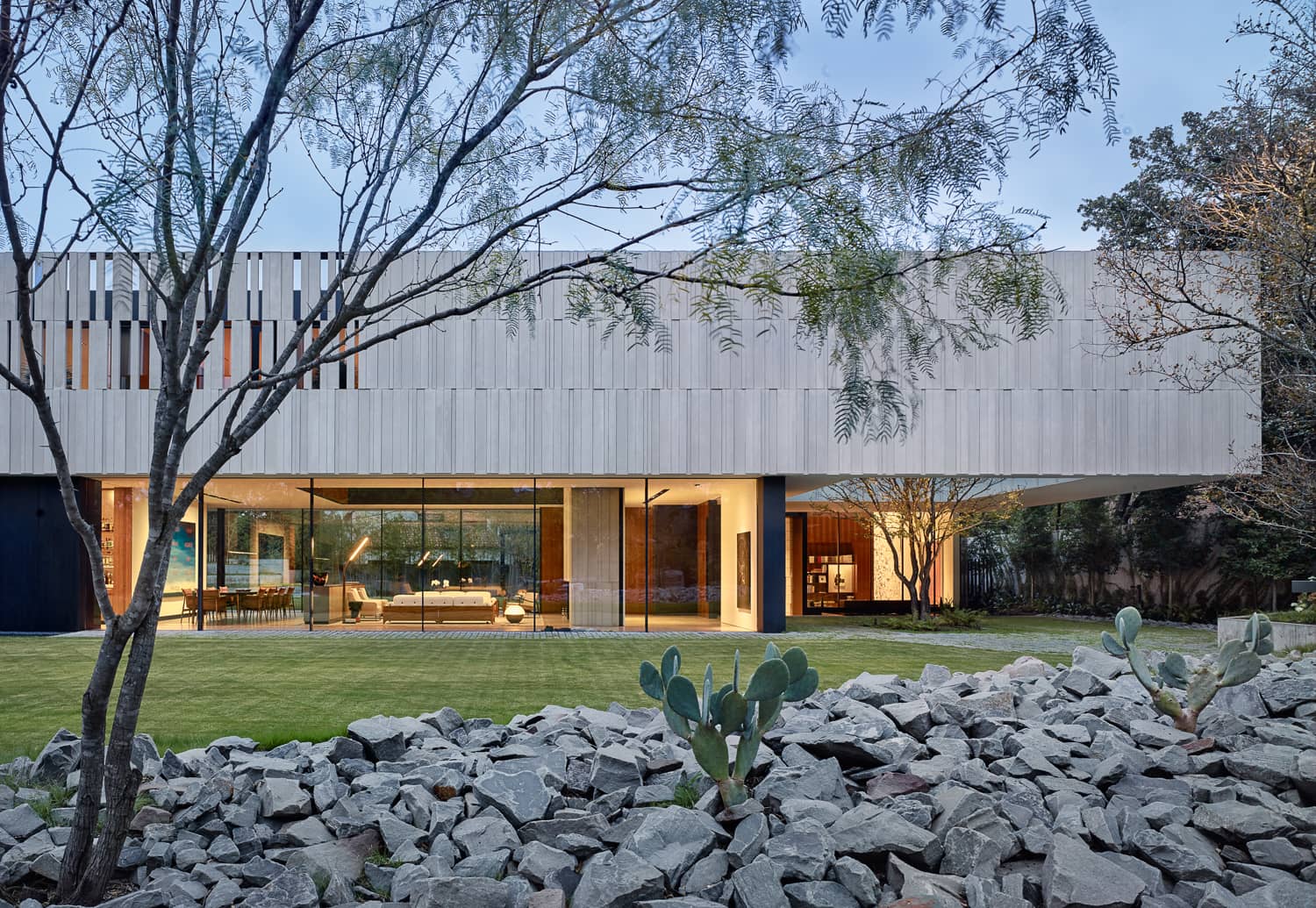 Modern architectural home with a sleek facade, large glass windows, and a landscaped yard featuring rocks, cacti, and trees.