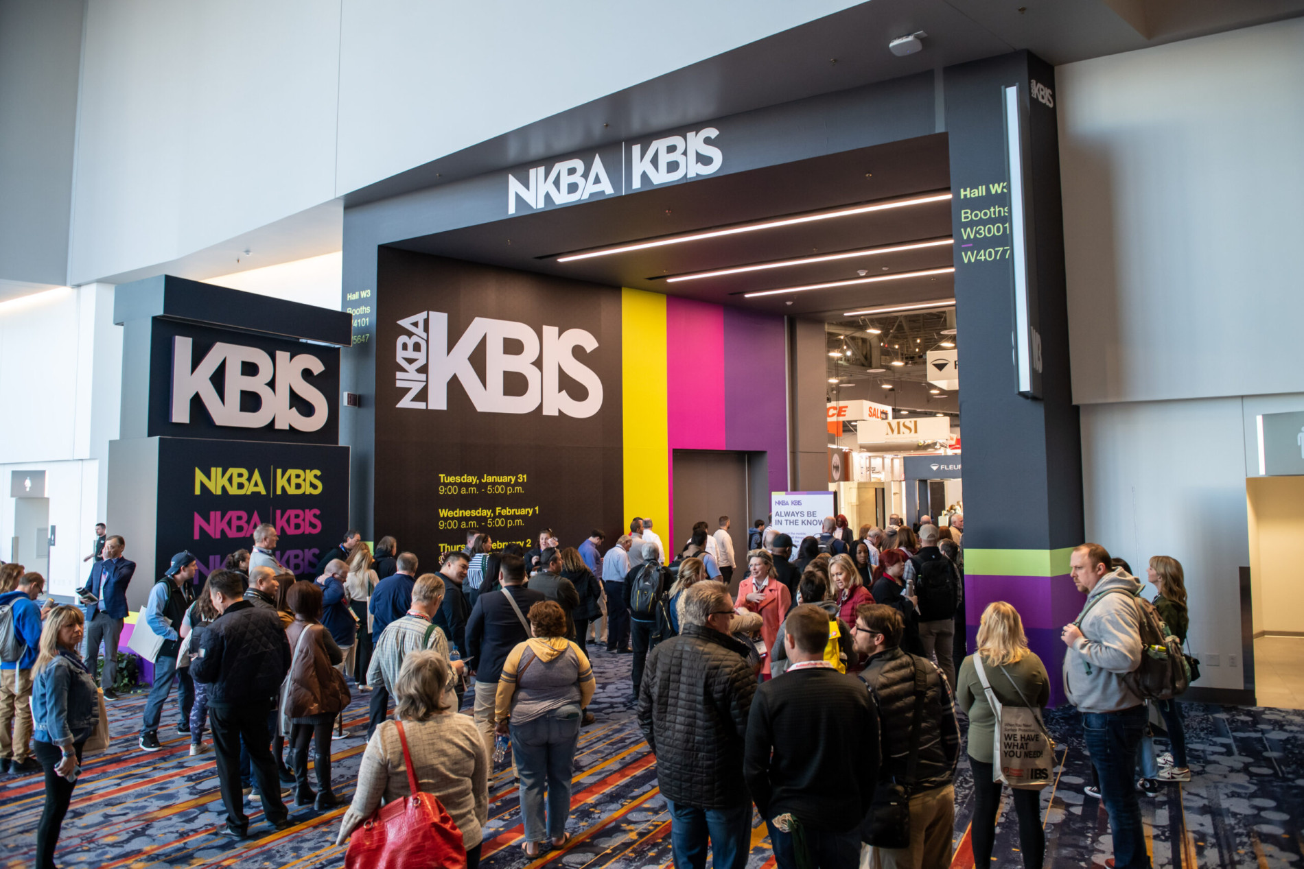 Crowds of attendees gather at the NKBA KBIS entrance, featuring tall signage with the event name and dates displayed on a modern, colorful design. Inside, exhibition booths and lights create an inviting atmosphere.