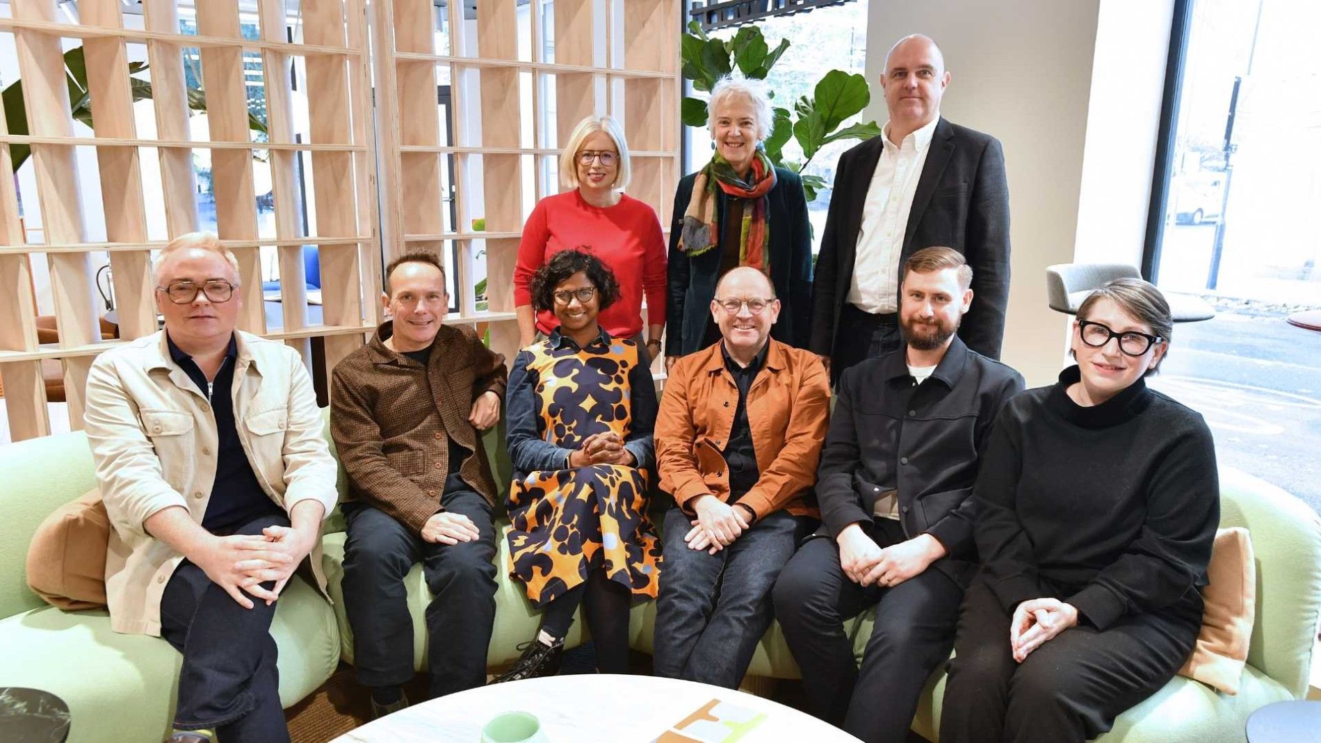 A group of nine people gathered in a modern indoor setting, seated on light green furniture or standing behind it. The group includes five men and four women, dressed in casual and professional attire, smiling at the camera. A wooden partition and indoor plants are visible in the background.