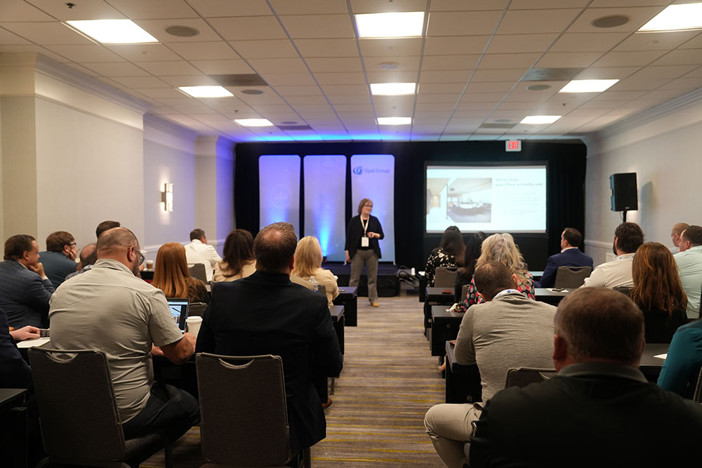 Speaker presenting to an engaged audience in a conference room setting, featuring a large projection screen and branded banners in the backgroun