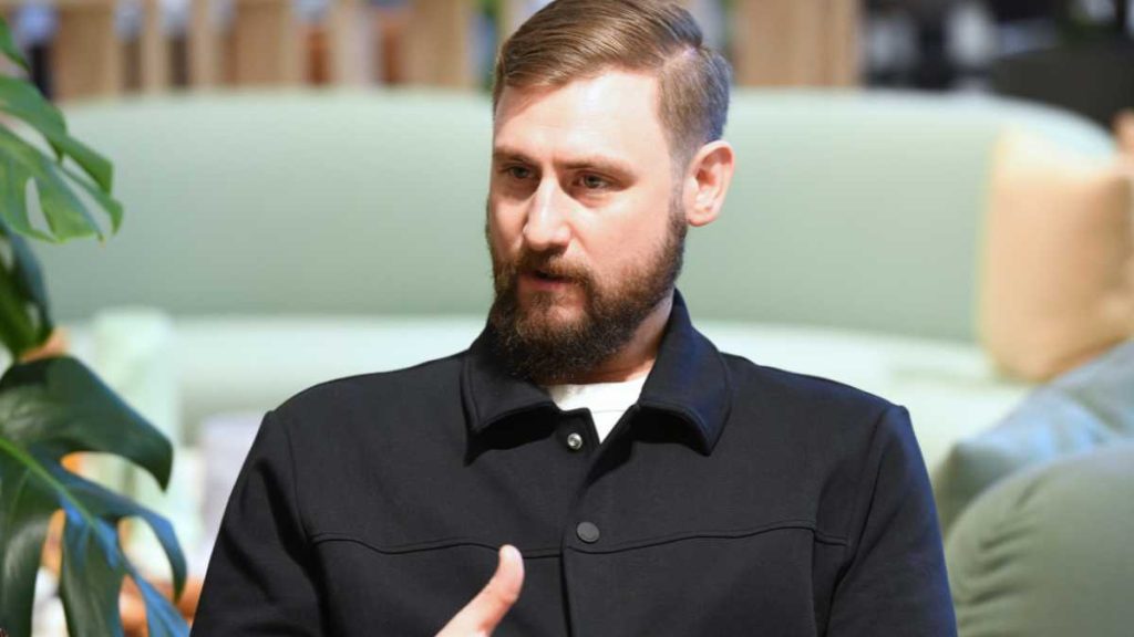 Man with a beard wearing a dark jacket, seated in a modern office space.