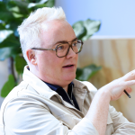 Man with blond hair and glasses gesturing while speaking in a discussion.