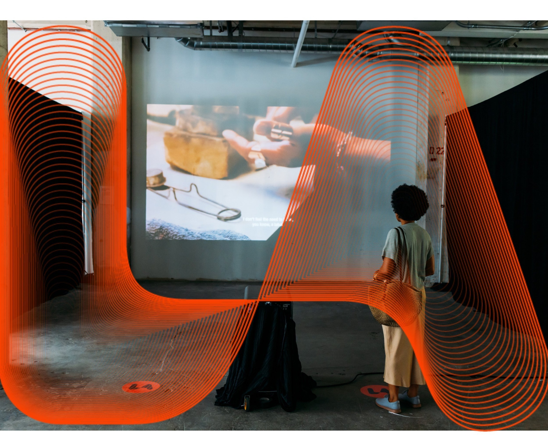 Person viewing an art installation with a projection of hands working on a tool, framed by a dynamic red geometric design in an industrial gallery space