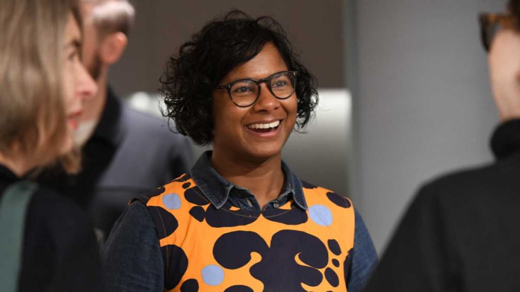 A smiling person with short, curly black hair and glasses, wearing a colorful orange and navy patterned top over a denim shirt, engaging with others in a casual setting.