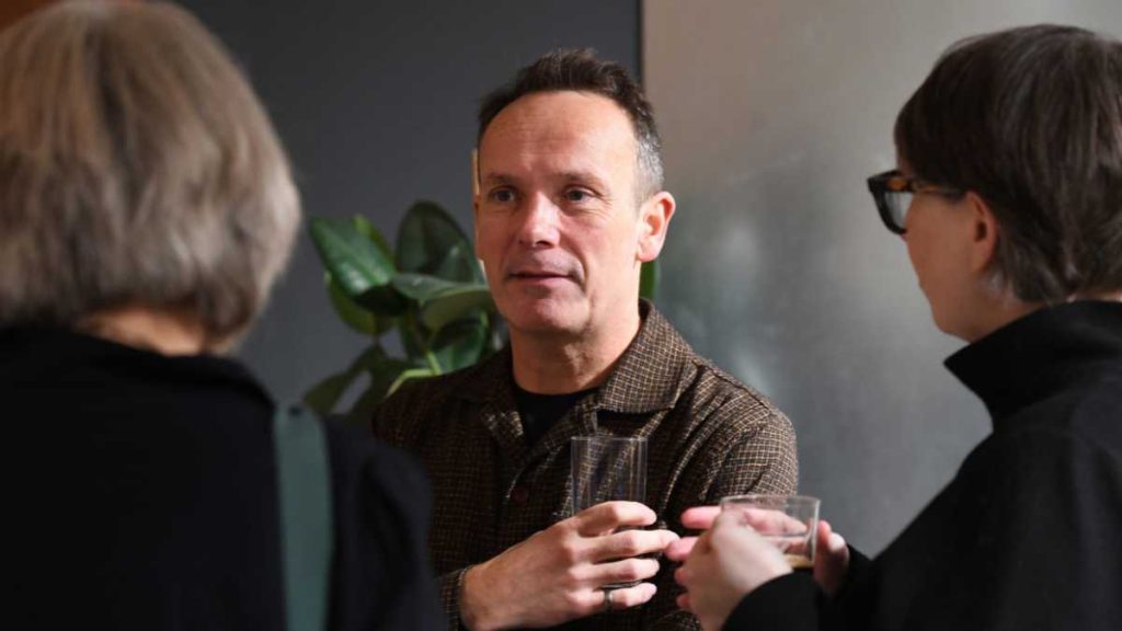 Man in a checked shirt holding a glass, engaging in conversation with two people in a social setting.