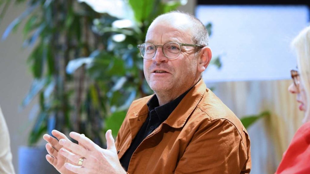 Man in glasses and an orange jacket speaking and gesturing in a meeting.