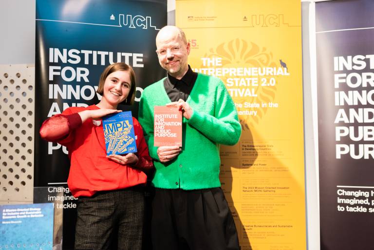 Two people stand in front of UCL banners, smiling and holding books related to innovation and public purpose. The woman wears a red sweater and black pants, while the man wears a green cardigan over black clothing.