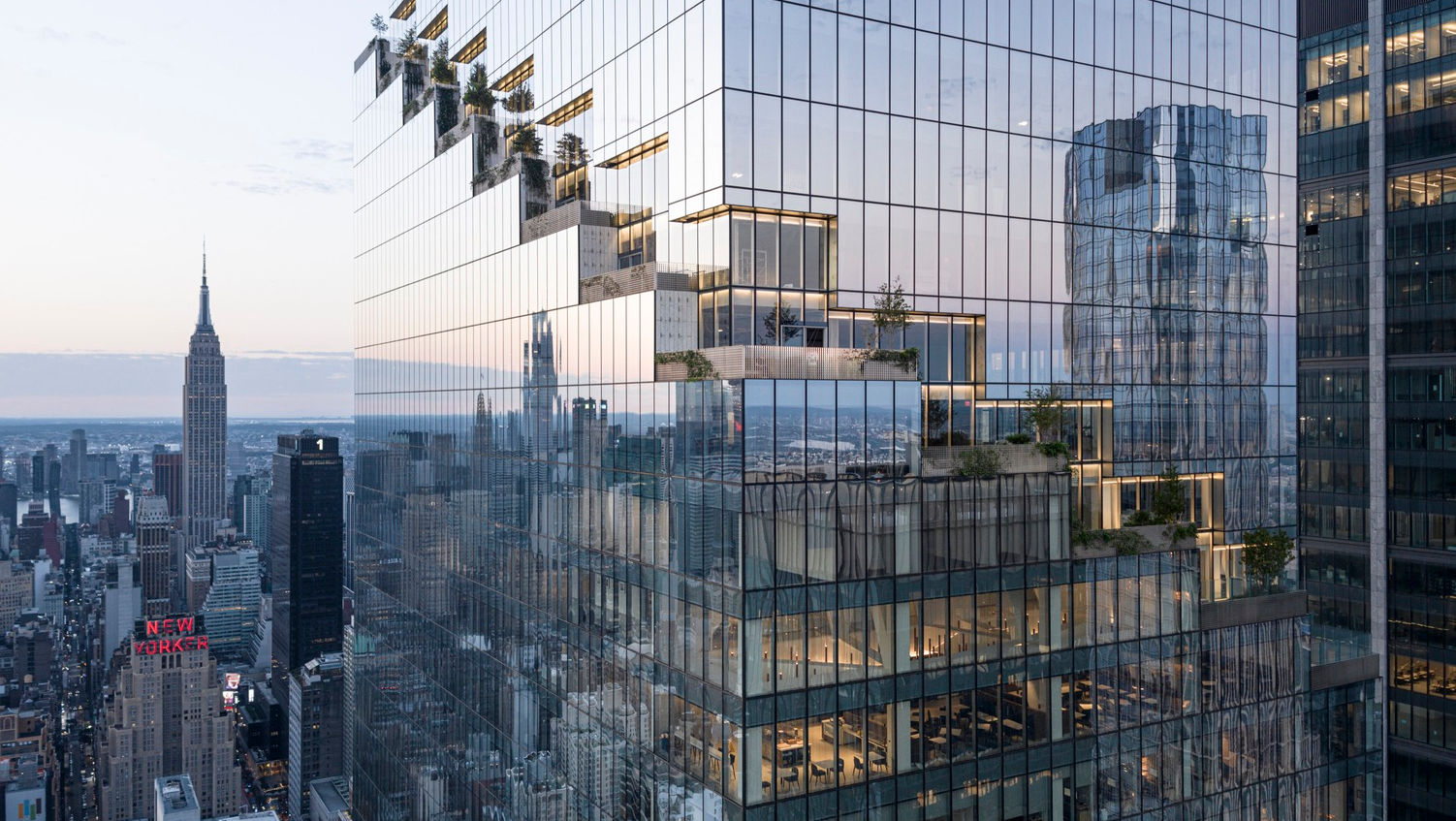 A modern glass skyscraper with cascading terraces and greenery overlooks the New York City skyline, including the Empire State Building at sunset.