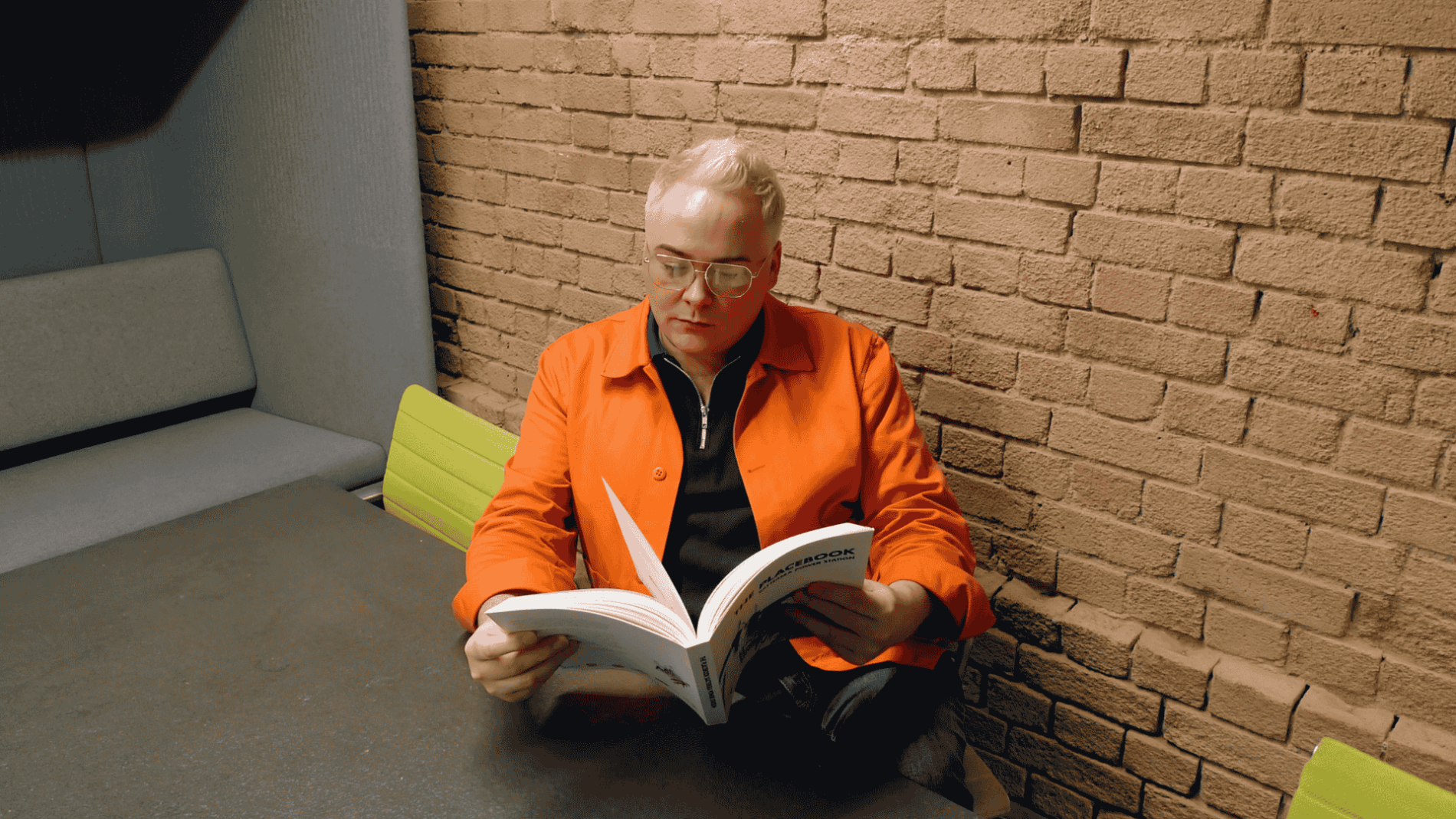 Seated man in orange shirt reading book in front of brick wall