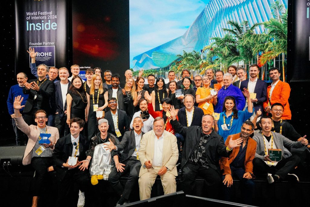 A large group of architects and designers celebrate at the World Architecture Festival 2024, holding awards and wearing event lanyards. They pose in front of a screen displaying a modern architectural design, with event branding in the background.