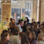 A networking event in a modern space with exposed brick walls and large windows. A diverse group of professionals, mostly women, are engaged in conversation, holding drinks. A woman in the foreground arranges champagne glasses on a wooden table