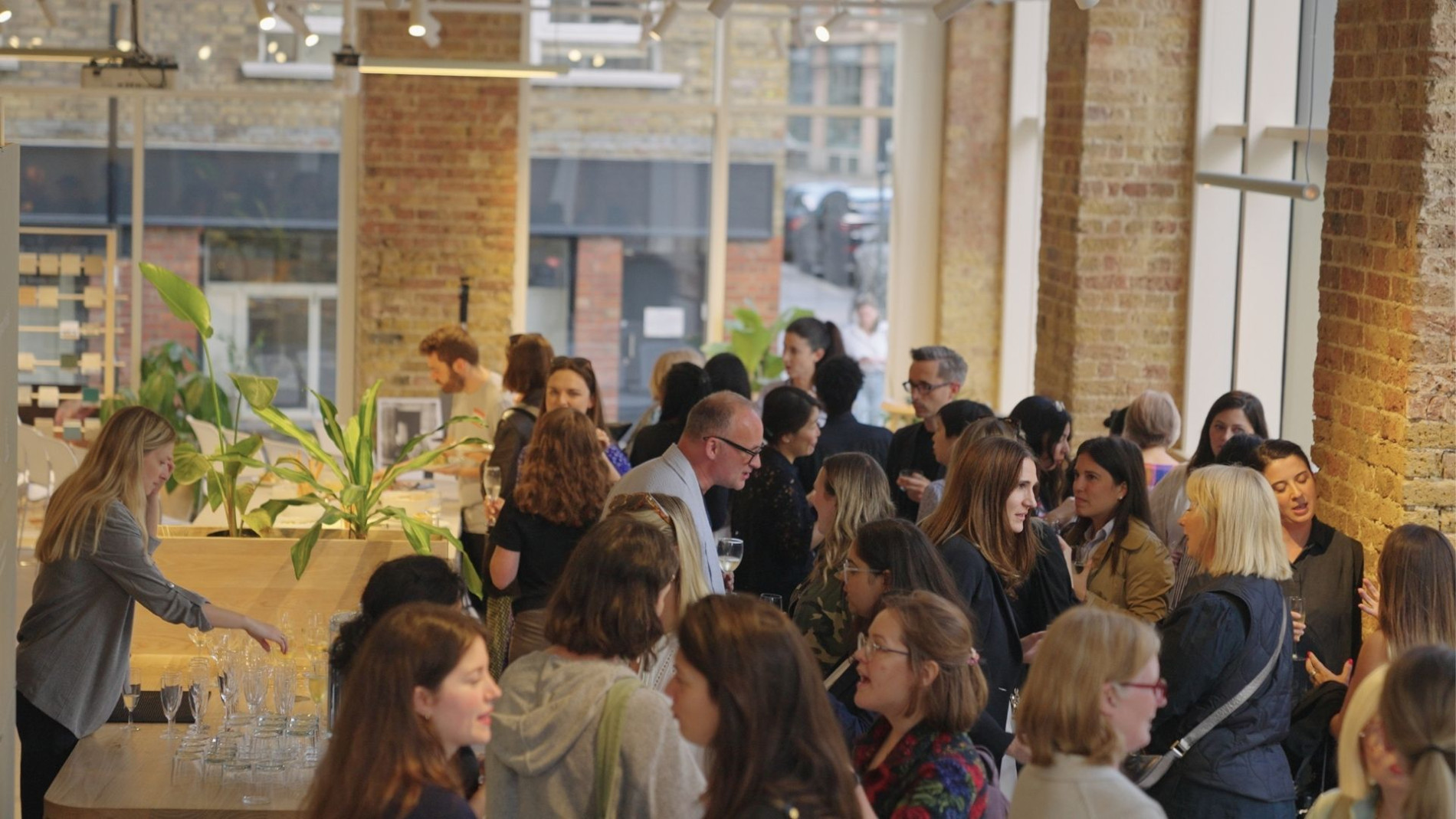 A networking event in a modern space with exposed brick walls and large windows. A diverse group of professionals, mostly women, are engaged in conversation, holding drinks. A woman in the foreground arranges champagne glasses on a wooden table