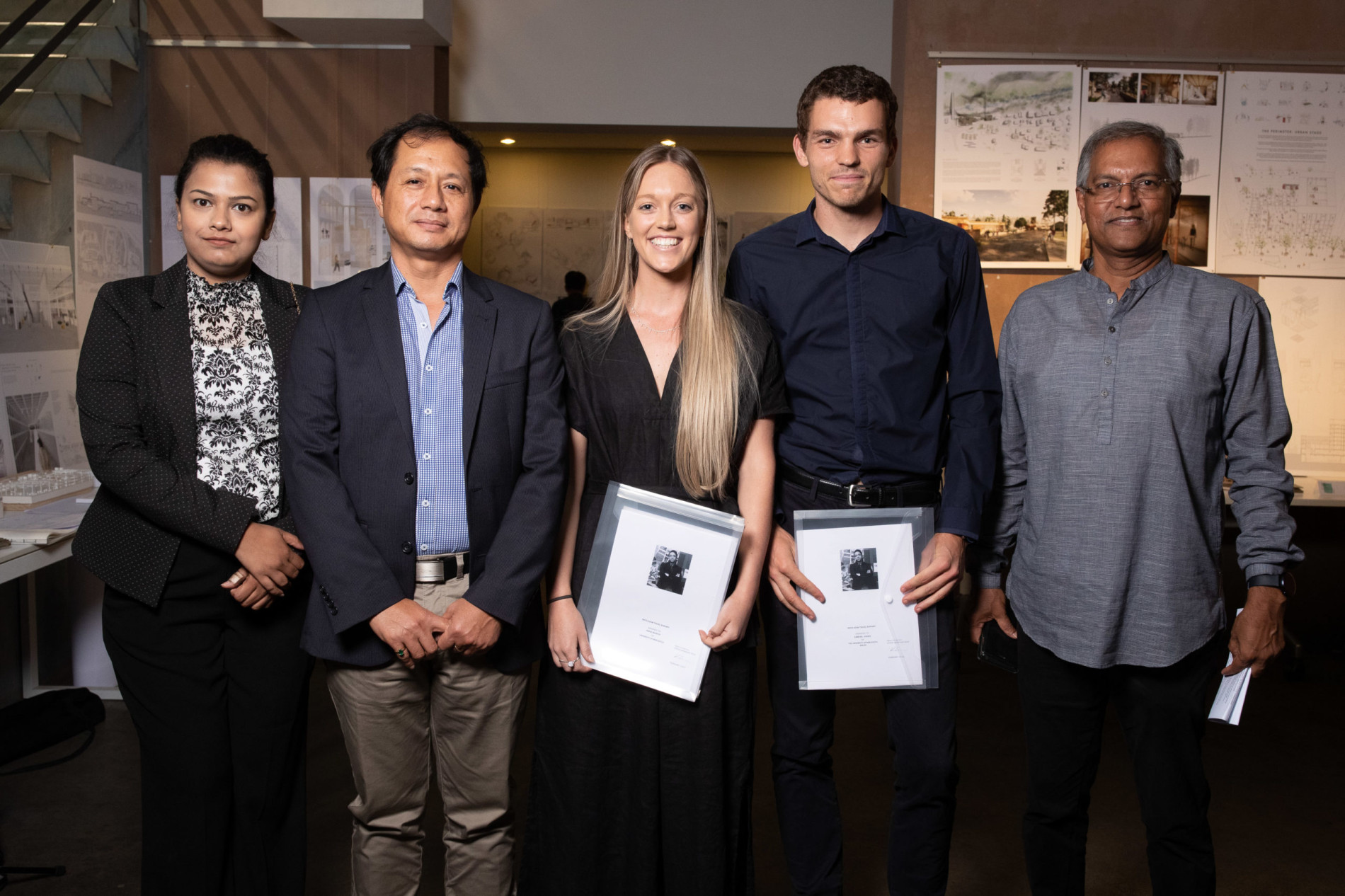 A group of five people posing at an architecture-related event. Two individuals in the center, a woman in a black dress and a man in a navy blue shirt, hold certificates in clear plastic folders. The other three, dressed in professional attire, stand on either side of them. The background features architectural drawings, models, and presentation boards on display. The setting is indoors with warm lighting.