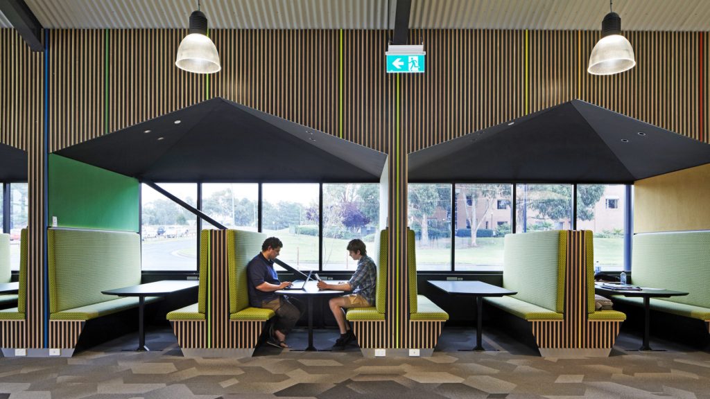 A modern workspace featuring semi-enclosed green fabric booths with black tables, designed for collaboration. The booths are accented by geometric wooden panelling and large windows that overlook a green landscape, offering a professional yet inviting environment for mentoring sessions.