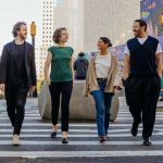 Four professionals walk together across a pedestrian crossing in New York City, engaged in conversation. They are dressed in a mix of smart and casual attire, with a backdrop of modern architecture and colourful street art.
