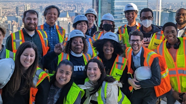 Group of diverse professionals wearing high-visibility vests and hard hats at a construction site with a city skyline in the background, smiling and posing for a photo.