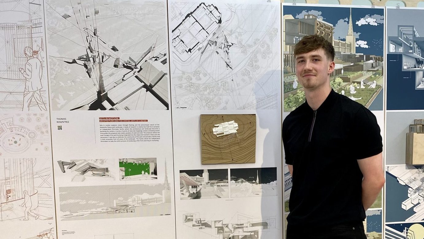 A young man stands in front of a display of architectural drawings and models mounted on vertical panels. He is dressed in a black shirt and trousers, with hands behind his back, smiling slightly. The display features detailed architectural plans, perspective renderings, and a small physical model. The setting appears to be a student exhibition or design review.